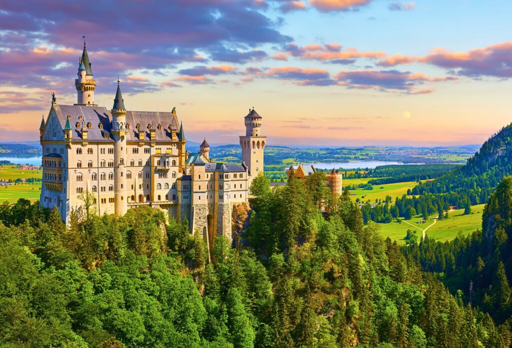 Castelo Neuschwanstein na Baviera, Alemanha, com floresta ao redor e céu colorido ao entardecer.