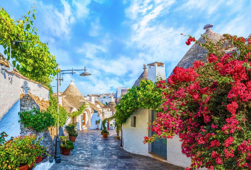 Rua florida de Alberobello com casas trulli, fachadas brancas e bougainvilleas coloridas na Puglia.